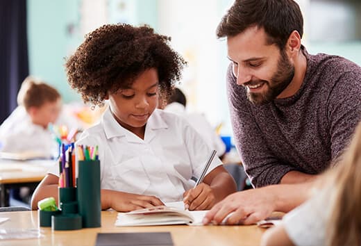 Candidate Sourcing A smiling teacher sitting with a young student and helping them with an assignment - education staffing
