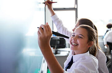 Young students in uniform, smiling and writing on a whiteboard - Teacher recruitment