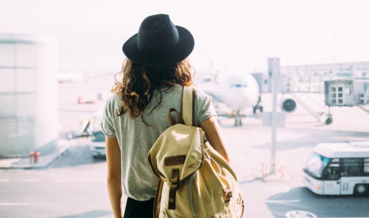 woman in a hat and wearing a backpack at the airport looking out at an airplane