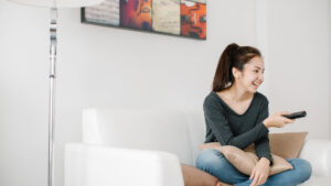 woman smiling and sitting on a white couch holding a remote facing the TV for ESL movies and TV shows for English language learners article