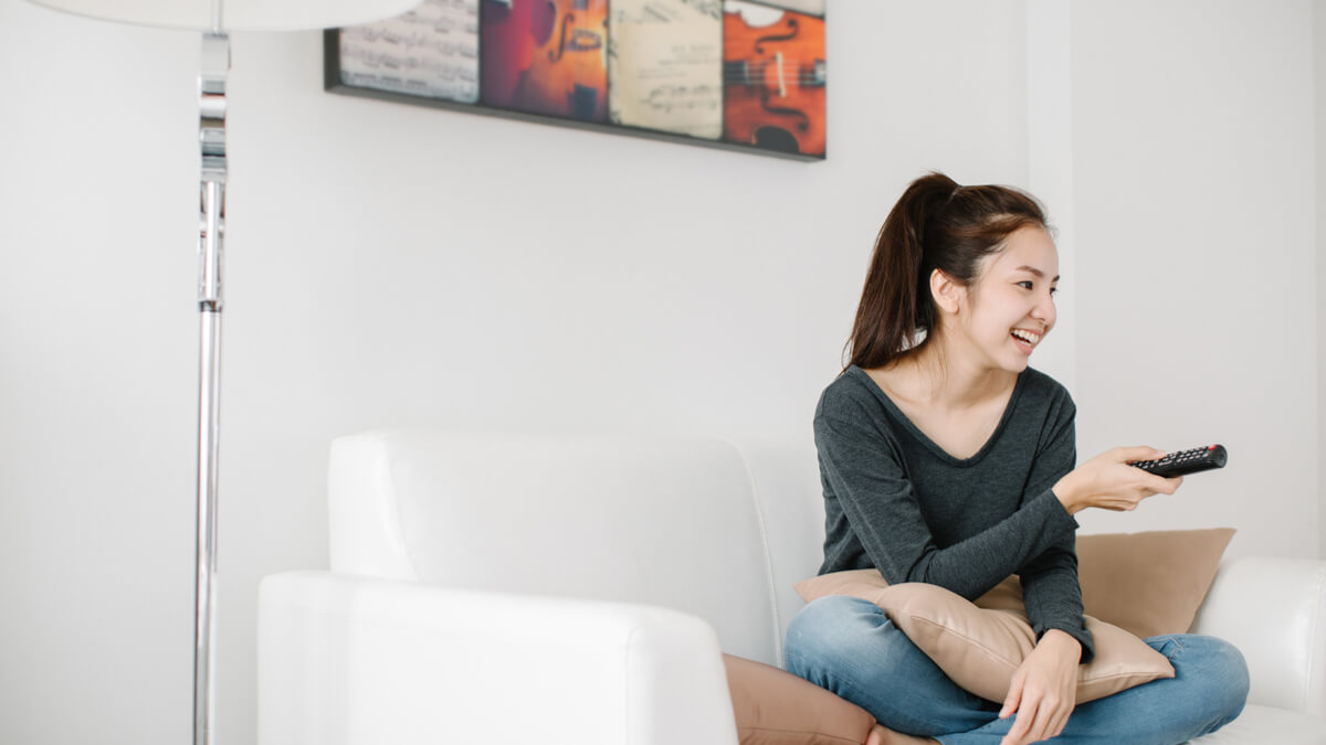 woman smiling and sitting on a white couch holding a remote facing the TV for ESL movies and TV shows for English language learners article