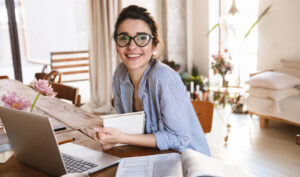 woman wearing glasses smiling in a sunny apartment and sitting in front of a laptop for esl resources for new teachers article
