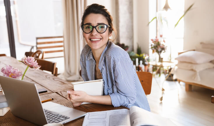woman wearing glasses smiling in a sunny apartment and sitting in front of a laptop for esl resources for new teachers article