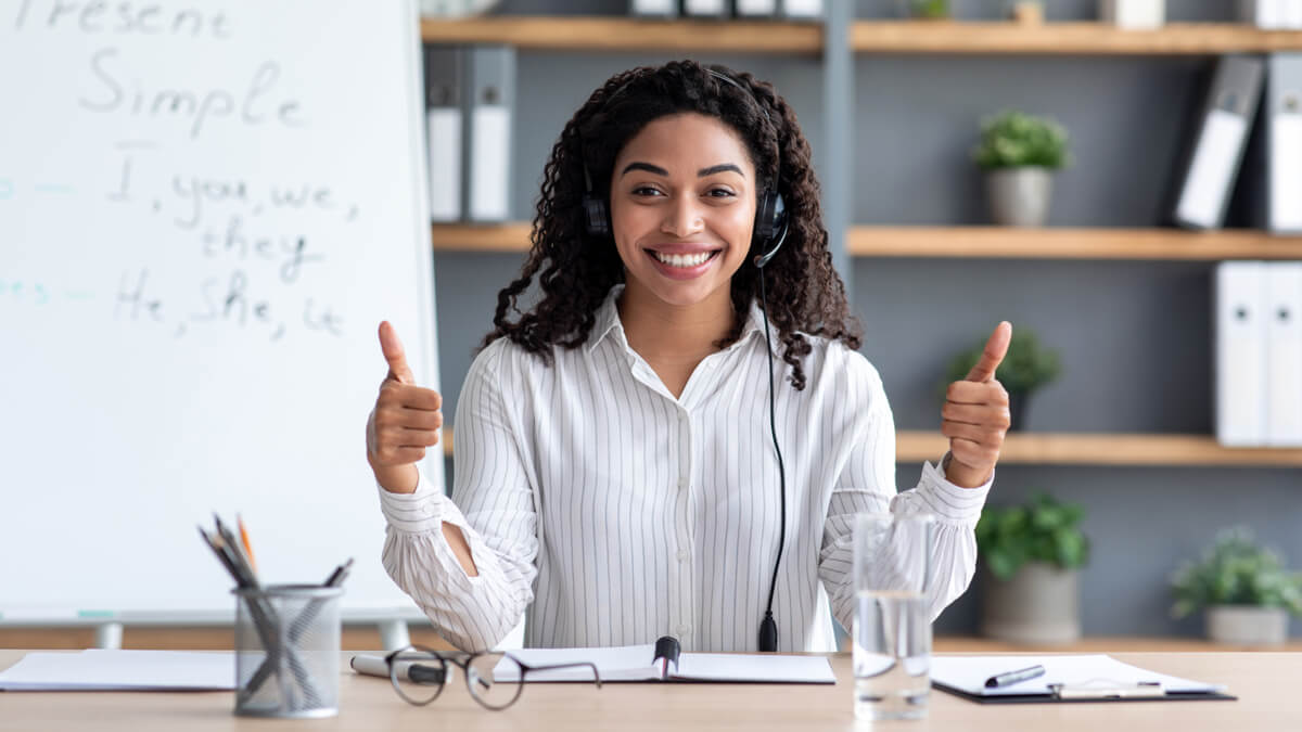 teacher sitting at a desk with thumbs up giving feedback in the online classroom for english language learners
